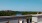 The Charley Apartments | A rooftop patio at Charley Apartments in Allston MA features five black cushioned chairs around a round fire pit table, decorative pillows, potted plants, and a scenic view of trees, the river, and distant buildings. | Apartments 