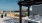 The Charley Apartments | A modern rooftop patio at Charley Apartments in Allston MA features a pergola, outdoor kitchen, grill, yellow chairs and tables, potted plants, and a large TV screen showing a soccer game against the city skyline backdrop. | Apart