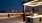 The Charley Apartments | A modern rooftop terrace at Charley Apartments in Allston MA features a lit pergola with a built-in grill, bar-height tables and chairs, city buildings in the background, and potted plants along a sleek railing. | Boston Waterfron