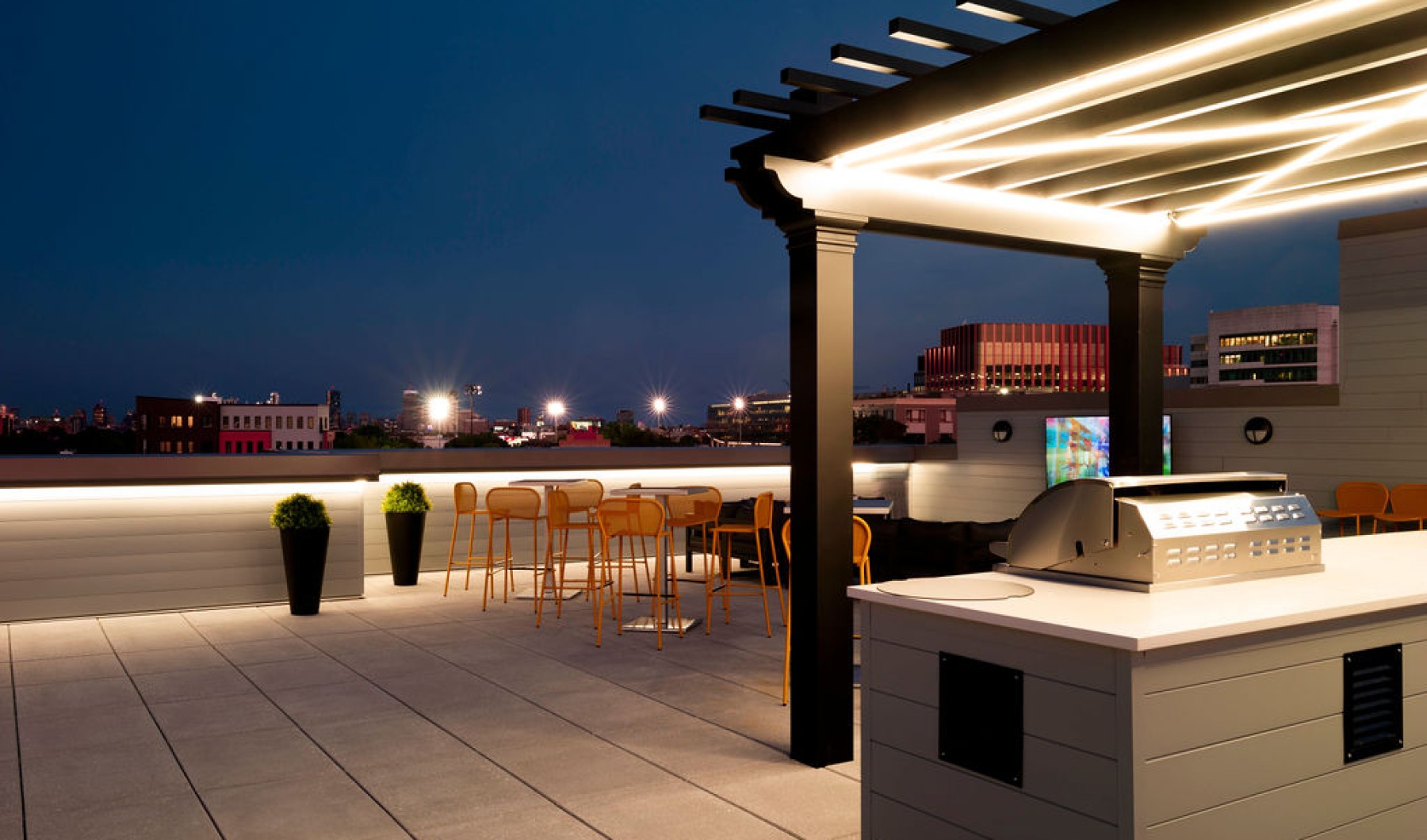 The Charley Apartments | A modern rooftop terrace at Charley Apartments in Allston MA features a lit pergola with a built-in grill, bar-height tables and chairs, city buildings in the background, and potted plants along a sleek railing. | Boston Waterfron